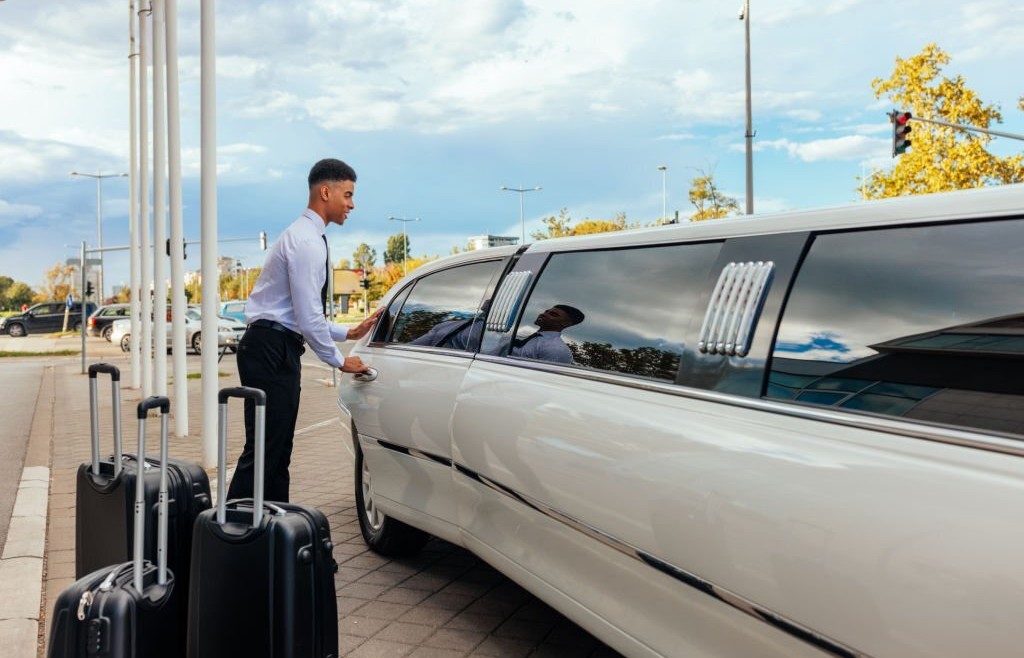 A young African American chauffeur is closing the door of the limousine after his clients entered the vehicle. There are some suitcases standing next to the car.